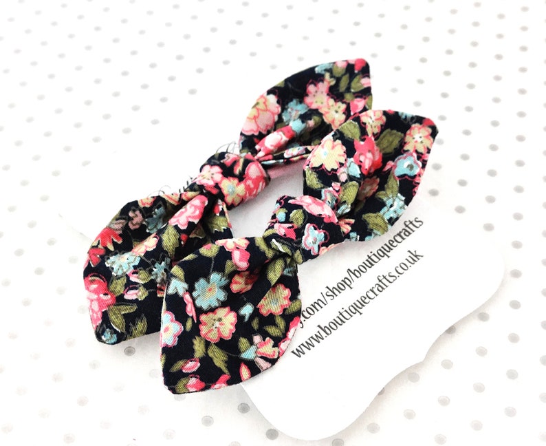 May include: Two black fabric hair bows with a floral pattern in pink, blue, and yellow. The bows are tied in a bunny ear style. A white tag with text is in the background.