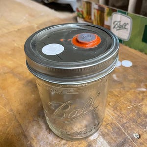 Puede incluir: Un tarro de vidrio transparente con una tapa plateada. La tapa tiene una pegatina blanca y un pequeño círculo naranja y gris en la parte superior. El tarro está etiquetado con "Ball" y tiene las palabras "wide mouth" impresas en la parte inferior.