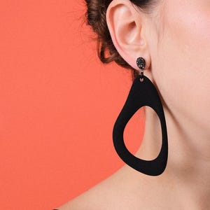 Può includere: Orecchini neri a goccia con un design ritagliato. Gli orecchini hanno un piccolo borchia rotonda nera nella parte superiore e una grande forma a goccia aperta. Gli orecchini sono mostrati su uno sfondo arancione.