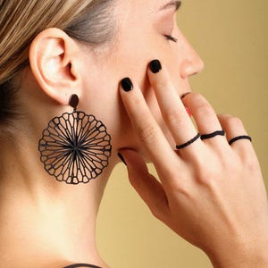 Può includere: Una donna con smalto nero e anelli neri alle dita. Indossa grandi orecchini neri con un design circolare e geometrico.
