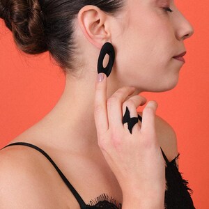 Puede incluir: Pendientes ovalados negros y un anillo a juego con dise&ntilde;o de rayo. La persona que lleva las joyas tiene el pelo oscuro recogido en un mo&ntilde;o. El fondo es de color naranja liso.