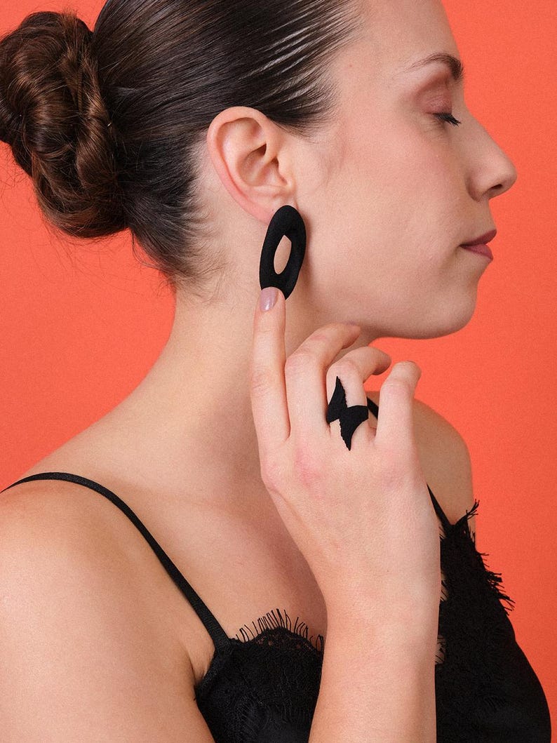 Puede incluir: Pendientes ovalados negros y un anillo a juego con dise&ntilde;o de rayo. La persona que lleva las joyas tiene el pelo oscuro recogido en un mo&ntilde;o. El fondo es de color naranja liso.