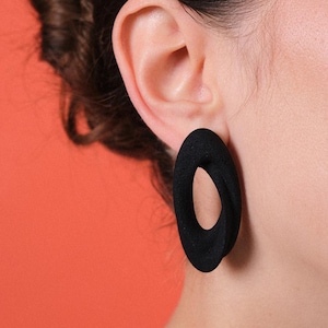 Puede incluir: Primer plano de una oreja con un pendiente negro en forma de óvalo. El pendiente tiene una superficie texturizada y un agujero en el centro. El fondo es de color naranja liso.