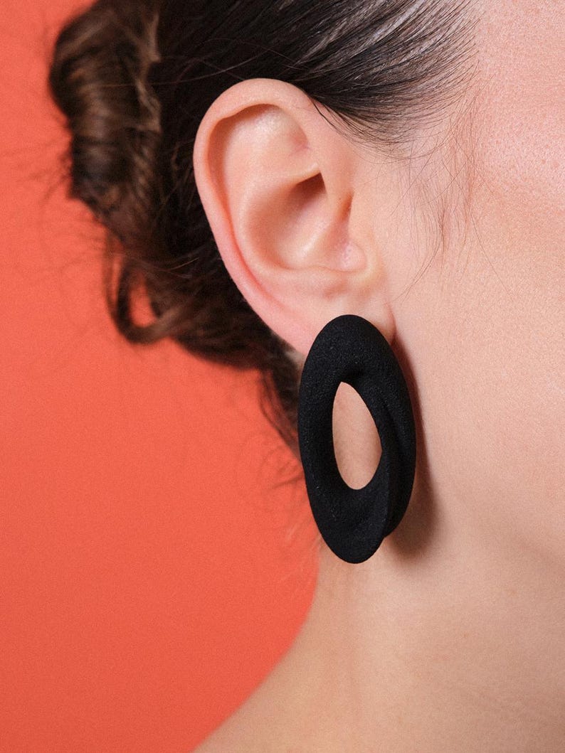 Puede incluir: Primer plano de una oreja con un pendiente negro en forma de &oacute;valo. El pendiente tiene una superficie texturizada y un agujero en el centro. El fondo es de color naranja liso.