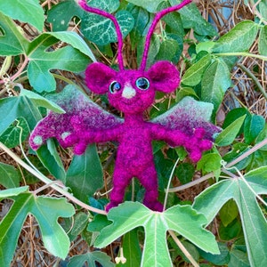May include: A small, purple, felt creature with large green eyes and long antennae. The creature has wings and is sitting in a bed of green leaves.