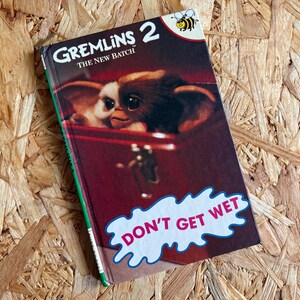 May include: A vintage copy of "Gremlins 2: The New Batch" on a wooden surface. The book cover features a close-up of a Gremlin and the text "DON'T GET WET". A small bee graphic is visible at the top.