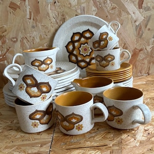 May include: A vintage ceramic dinner service with speckled white mugs, saucers, plates, and serving dishes. The set features a brown and orange floral design. The mugs and serving dishes have a brown interior.