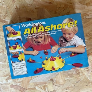 May include: A retro board game box for "All Ashore!" by Waddingtons, showing children playing. The game features a yellow flower-shaped board, small wooden figures, and red boats. The box states the game is for 2-4 players, suitable for ages 4 and up.