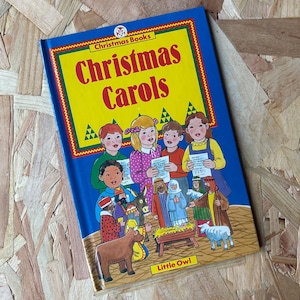 May include: A children's book titled "Christmas Carols" with a blue cover and a yellow background. The cover features illustrations of children singing carols and a nativity scene. The book is from "Christmas Books" by Little Owl.