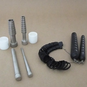 May include: A collection of jewelry tools, including ring sizers, mandrels, and sizing gauges. The set includes black ring sizers, two metal mandrels, two white cylindrical objects, and two metal tools. The tools are arranged on a neutral background.