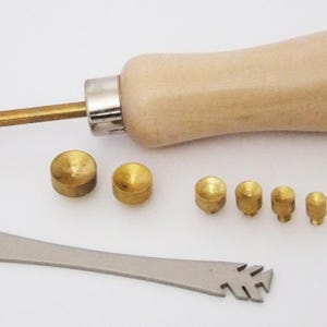 May include: A leathercraft tool set featuring a wooden handle, brass components, and a metal cutting tool. The set includes various brass tips and a metal blade, ideal for leatherworking projects. The tools are arranged on a white surface.