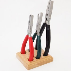 Pode incluir: Um conjunto de alicates para joias com cabos vermelhos, verdes e pretos, exibidos em um suporte de madeira. Os alicates têm cabeças de metal prateado e são projetados para trabalhos artesanais detalhados. O suporte de madeira tem quatro orifícios.