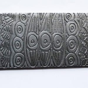 May include: A rectangular, gray metal object with an intricate, embossed design. The pattern features a symmetrical arrangement of spirals, circles, and linear elements, creating a textured surface. The object appears to be a decorative element or a piece of art.