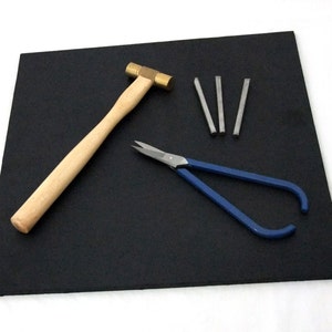 Puede incluir: Una estera de goma negra con un martillo de lat&oacute;n, alicates azules y cinco varillas de metal plateado.