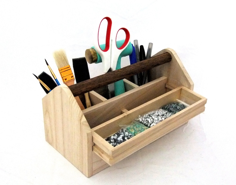 May include: A wooden toolbox with a brown handle, divided into compartments. It contains various tools, including paintbrushes, scissors, and a hammer. The toolbox has a pull-out drawer filled with silver and green beads.