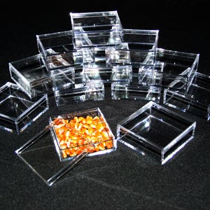 May include: A collection of clear, square acrylic boxes, some stacked, others open or closed. One box contains golden-brown kernels. The boxes are arranged on a dark surface, creating a display of storage containers.