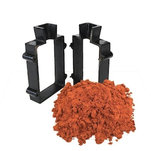 May include: Two black metal molds with a rectangular shape and a pile of reddish-brown powder. The molds have a unique design with a top and side extensions. The powder appears to be finely ground.