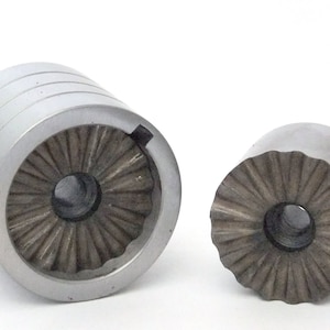 Op de afbeelding: Twee metalen koekjespersmatrijzen, een met een grotere diameter dan de andere. Beide matrijzen hebben een decoratief, gegolfd ontwerp rond het middelste gat.
