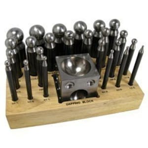 May include: A set of 25 metal dapping punches and a dapping block, organized in a wooden holder. The punches are various sizes and shapes, including round, square, and pointed.
