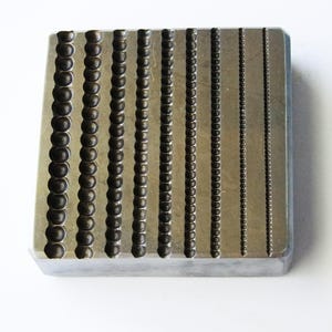 May include: A rectangular metal bead forming block with ten vertical channels of varying bead sizes. The beads are dark and rounded, and the block has a silver finish. This tool is used for jewelry making and crafting.