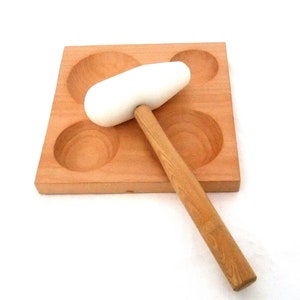 Könnte beinhalten: Ein weißer Holzhammer mit einem Holzstiel liegt auf einem Holzbrett mit vier runden Vertiefungen.