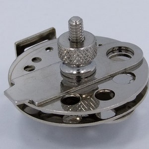 Op de afbeelding: Een zilverkleurig metalen gereedschap met een ronde basis en een schroef met schroefdraad. De basis heeft meerdere gaten van verschillende groottes en een klein, plat, rechthoekig stuk dat aan de zijkant uitsteekt.