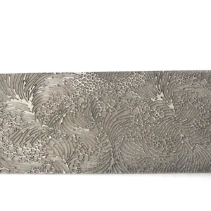 Peut inclure: Une pièce décorative rectangulaire argentée avec un motif texturé complexe. Le design présente un mélange de lignes ondulées et de petites formes circulaires, créant un effet artistique détaillé. La pièce est posée sur un fond blanc.