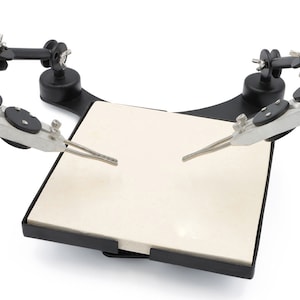 May include: A black and silver soldering station with adjustable arms and clamps. The station features a square, off-white ceramic base, ideal for holding small electronic components during soldering or repair work. The arms are articulated for flexible positioning.