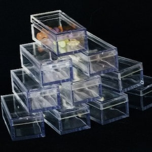 Peut inclure: Une pile de boîtes en plastique transparentes et rectangulaires avec des couvercles à charnière. Certaines boîtes contiennent de petits objets colorés. Les boîtes sont disposées en forme de pyramide sur fond noir.