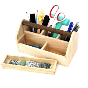 May include: A wooden tool box with a dark brown handle, divided into compartments, filled with various tools, including paintbrushes, scissors, pens, and a hammer. A separate wooden tray with a clear plastic bag filled with small silver and green beads sits in front of the box.