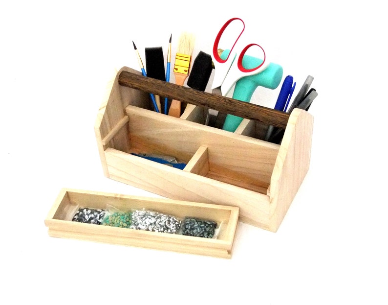 May include: A wooden tool box with a dark brown handle, divided into compartments, filled with various tools, including paintbrushes, scissors, pens, and a hammer. A separate wooden tray with a clear plastic bag filled with small silver and green beads sits in front of the box.