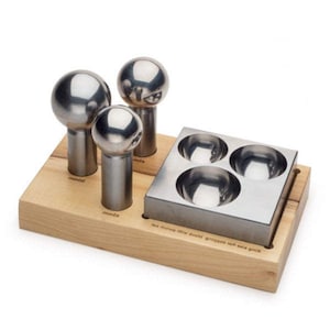 Peut inclure: Un ensemble d'outils de fabrication de bijoux comprenant trois outils en métal poli en forme de champignon et un bloc de formage en métal avec trois dépressions concaves. Les outils sont posés sur une base en bois clair.