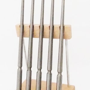 Puede incluir: Un juego de seis herramientas metálicas con una base de madera. Las herramientas son de color plateado, cilíndricas y tienen una base ligeramente más ancha. La base de madera es rectangular y de color claro. Las herramientas están dispuestas verticalmente.