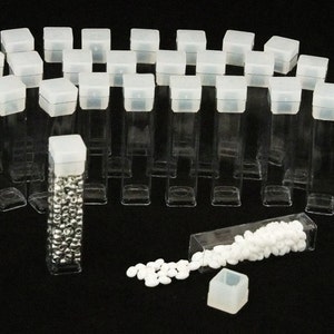 以下が含まれることがあります： 白い蓋が付いた透明なプラスチック容器が25個セットになっています。容器は半円形に配置されています。1つの容器には小さな銀色のビーズが入っています。もう1つの容器には白いビーズが入っており、黒い表面にこぼれています。