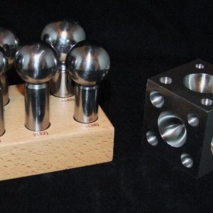 Puede incluir: Conjunto de cinco herramientas de metal en forma de bola con mangos, dispuestas en un soporte de madera. Las herramientas están etiquetadas como 1 (28), 2 (32), 3 (35), 4 (40) y 5 (50). También se muestra un cubo de metal con cinco depresiones redondas.