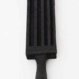 May include: A black cast iron ingot mold featuring a rectangular body with four parallel vertical channels and a short, rounded handle extending from the bottom. The surface has a textured, matte finish with a solid, heavy appearance.
