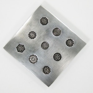 Könnte beinhalten: Eine quadratische, silberfarbene Metallplatte mit acht geprägten floralen Designs. Die Designs variieren im Stil, einige ähneln Gänseblümchen und andere komplexeren Mustern. Die Platte hat eine gebürstete Metalloberfläche und ist vor einem weißen Hintergrund platziert.
