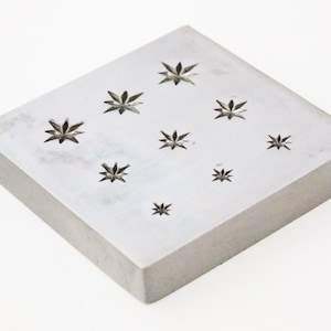 May include: A square metal block with multiple star-shaped cutouts. The block is a tool for creating impressions in metal.