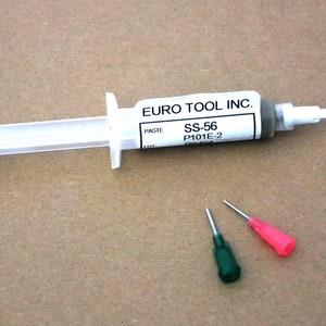 Könnte beinhalten: Eine weiße Spritze mit einer silbernen Spitze, gefüllt mit einer grauen Paste. Die Spritze trägt ein Etikett mit der Aufschrift "EURO TOOL INC. PASTE SS-56 P101E-2". Zwei Nadeln sind im Lieferumfang enthalten, eine grüne und eine rote.