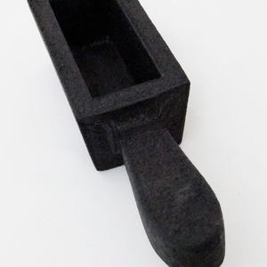 May include: A black rectangular mould with a textured surface and a short, rounded handle extending from one side. The object features a deep, hollow centre and a solid, matte finish, presenting a simple, functional design for casting or shaping.