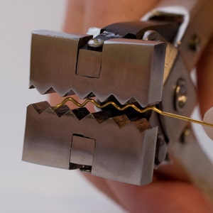 Op de afbeelding: Close-up van een metalen draadcrimptang met een gouden draad die wordt gecrimpt. De tang heeft een gekartelde bek en een hendel.