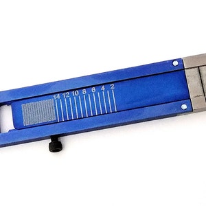 May include: A blue metal ruler with a scale marked in inches from 2 to 14. The ruler has a small hole on the left side and a screw on the bottom.