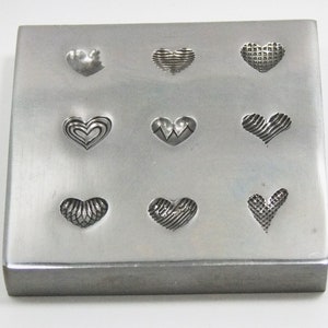 May include: A metal stamp with nine different heart-shaped designs. The stamp is made of silver metal and has a rectangular shape.