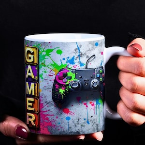 Puede incluir: Taza de cerámica blanca con un diseño de salpicaduras de pintura de colores y un mando de videojuego negro. La palabra "GAMER" está escrita verticalmente en letras de bloque de colores en el lado izquierdo de la taza.
