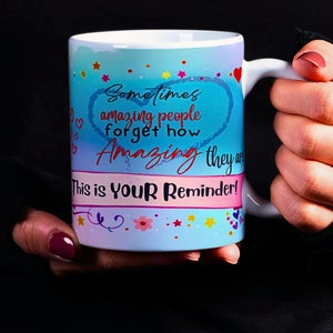 May include: A white ceramic mug with a blue and pink design. The mug has a heart shape with the text "Sometimes amazing people forget how Amazing they are. This is Your Reminder!"