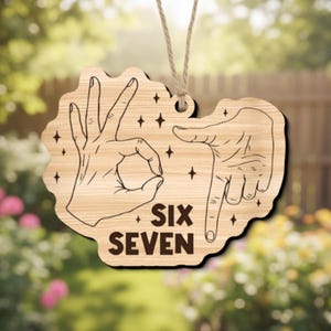 May include: Wooden sign with a light wood grain finish, featuring hand gestures for the numbers six and seven. The words "SIX SEVEN" are printed in a bold, dark font. The sign is suspended by a natural twine string, with star-shaped accents.