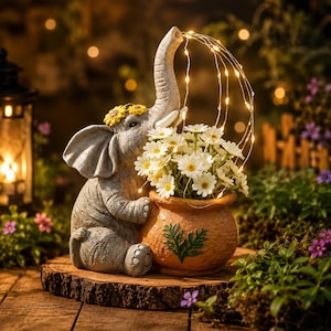 May include: A gray elephant figurine with a yellow sunflower crown, holding a textured orange pot filled with white daisy flowers. A string of thin, glowing fairy lights extends from the elephant's trunk, arching over the flowers.