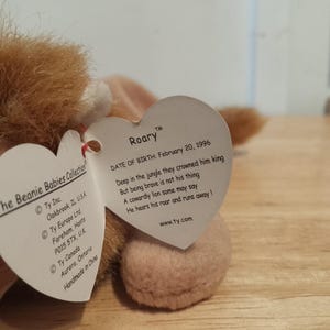 May include: A plush lion toy from The Beanie Babies Collection, featuring a brown mane and a tan body. The toy has a heart-shaped tag with the name "Roary" and a poem. The tag also includes the date of birth: February 20, 1996.