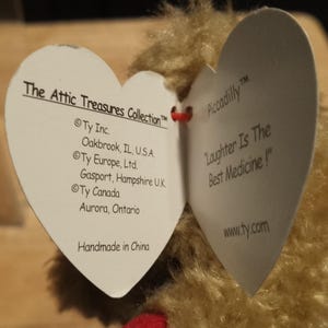 May include: Close-up of a plush toy with two heart-shaped tags. One tag reads "The Attic Treasures Collection" and lists locations. The other tag says "Piccadilly" and "Laughter Is The Best Medicine!" The toy is handmade in China.