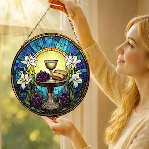 May include: A round stained glass window panel featuring a chalice, bread, and grapes on a table, surrounded by white lilies and blue and yellow stained glass. The panel is held by a chain.